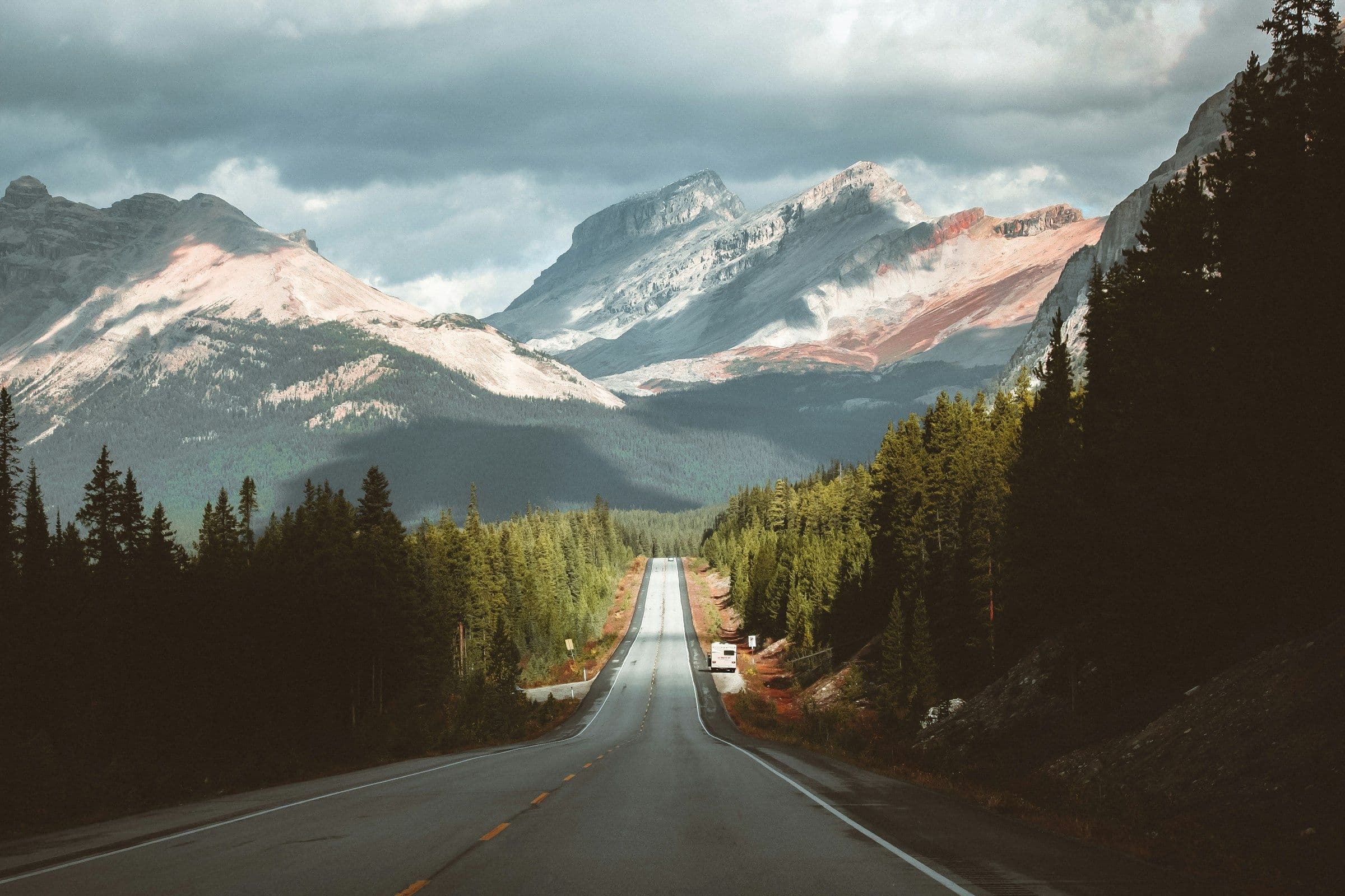 An empty road running between pine forest towards the mountains of Alberta, Canada.
