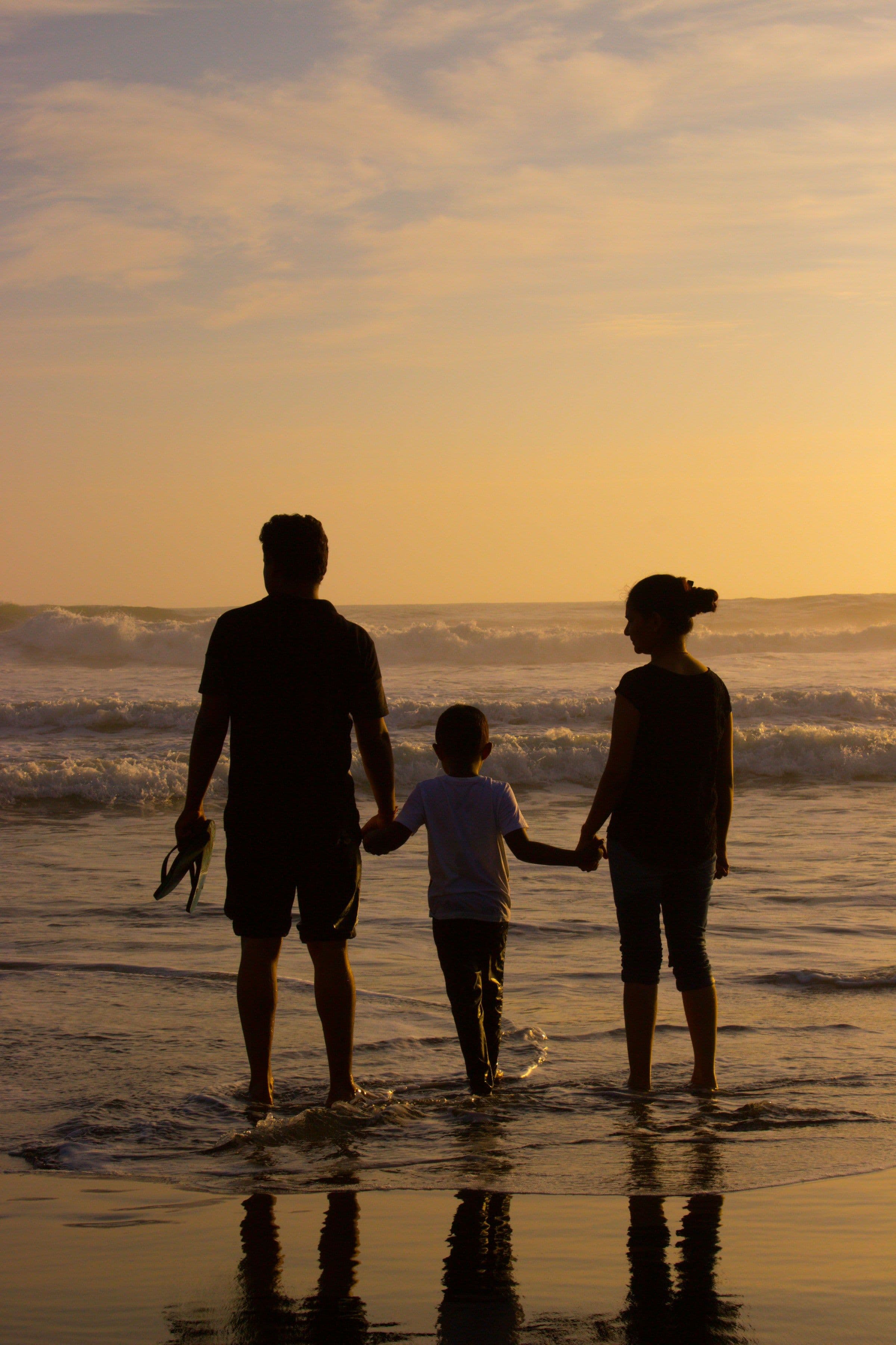 A family of three in silhouette, paddling at the water’s edge at sunset.