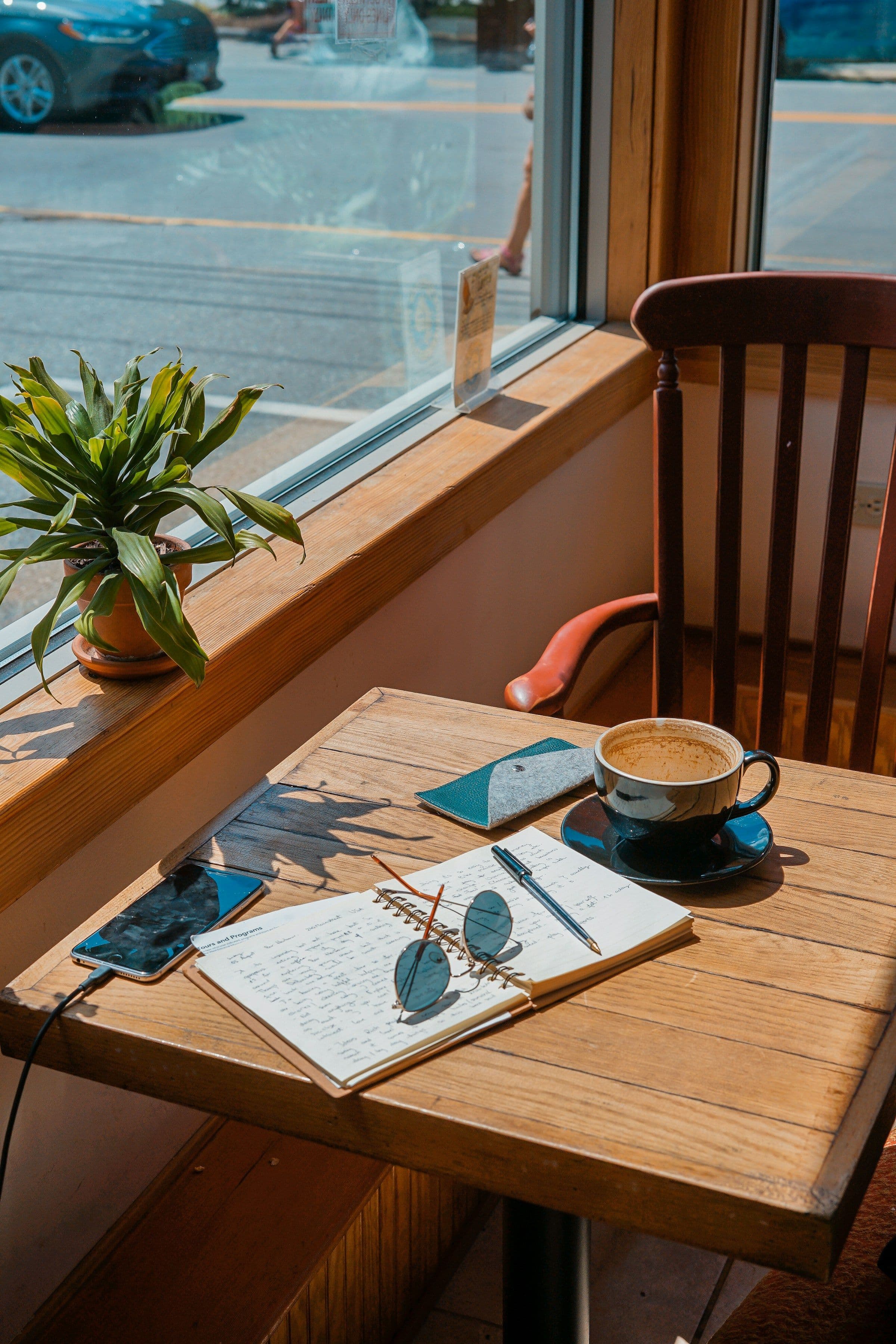 A cup of coffee, a notebook and sunglasses on a wooden table beside a café window.