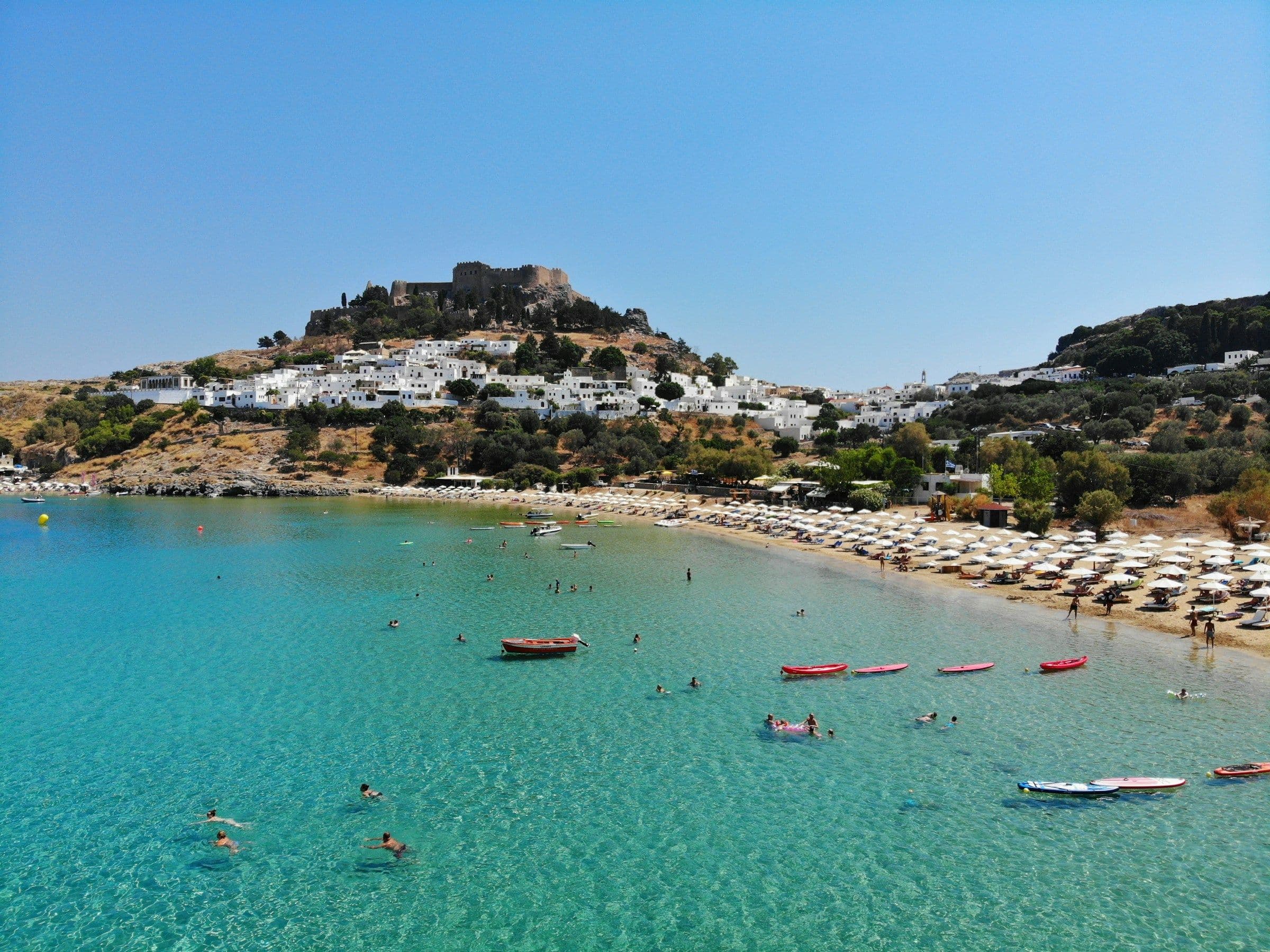 The white town of Lindos and its hilltop acropolis above a bay with a beach of sunshades, on Rhodes, Greece.