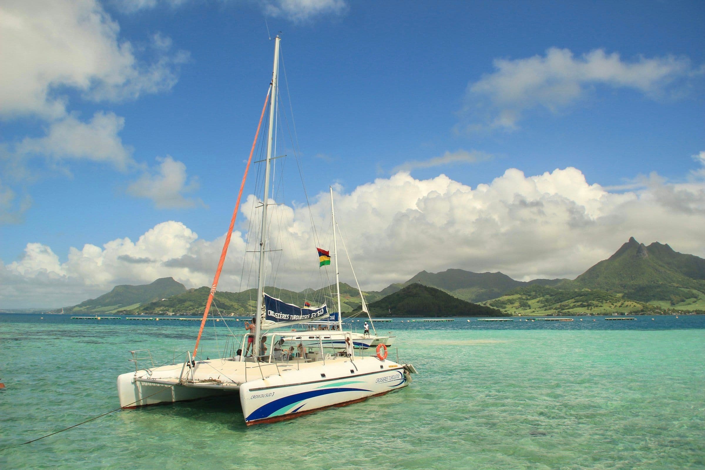 A catamaran at anchor in a shallow lagoon in Mauritius, with green mountains behind.