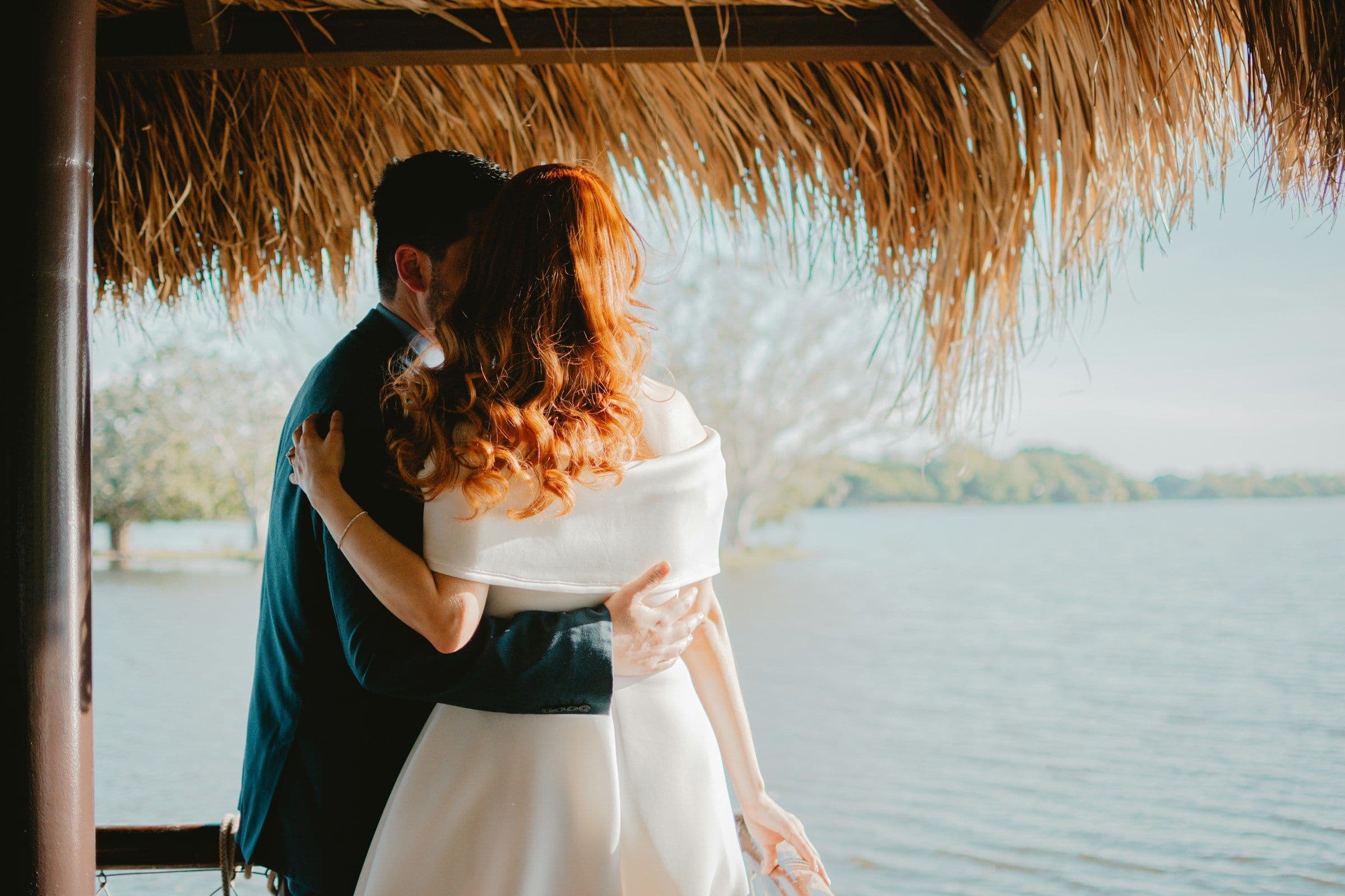 A couple embracing beneath a thatched roof, looking out over calm water.