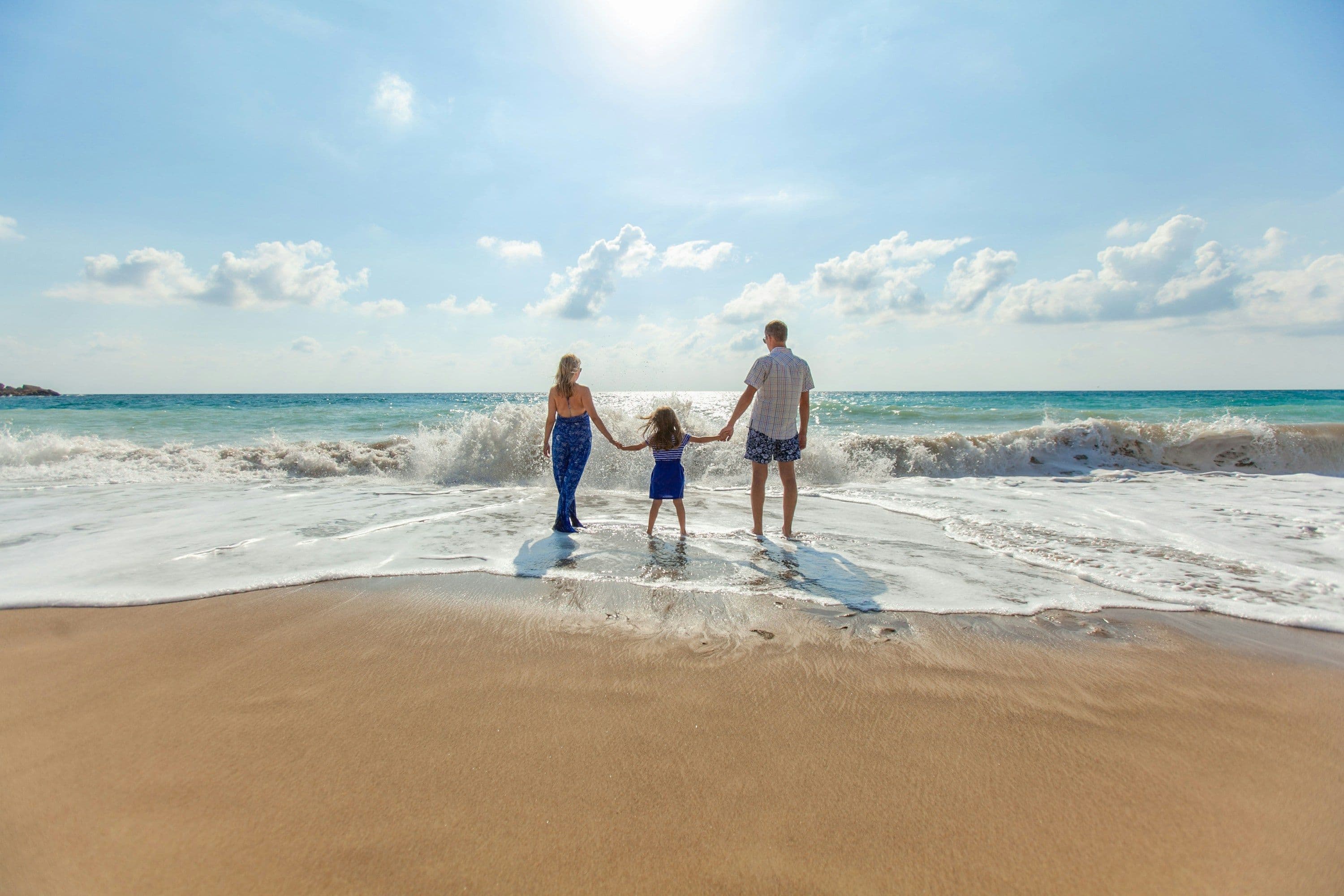 Two adults and a child holding hands at the edge of the surf on a sandy beach in Paphos, Cyprus, seen from behind.