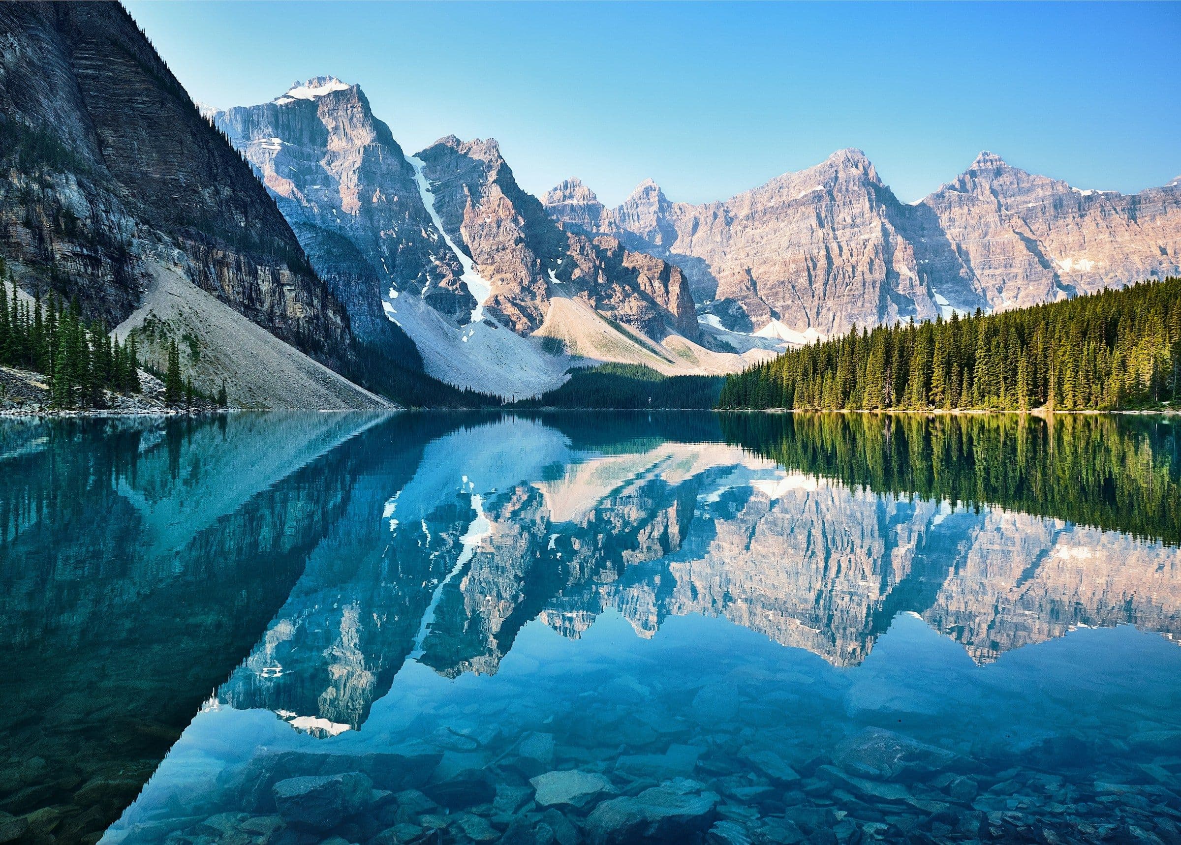 Snow-streaked mountain peaks and pine forest reflected in the still, clear water of Moraine Lake, Canada
