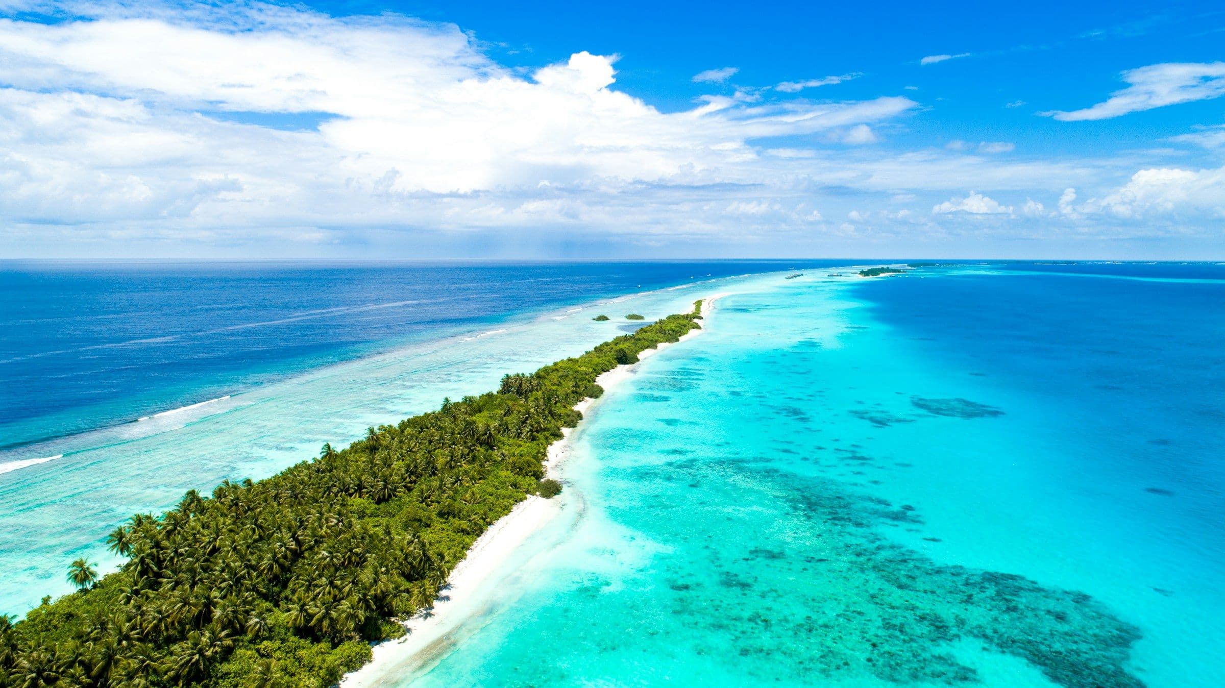 A long, narrow palm-covered island between deep blue sea and a turquoise lagoon in the Maldives, seen from the air.