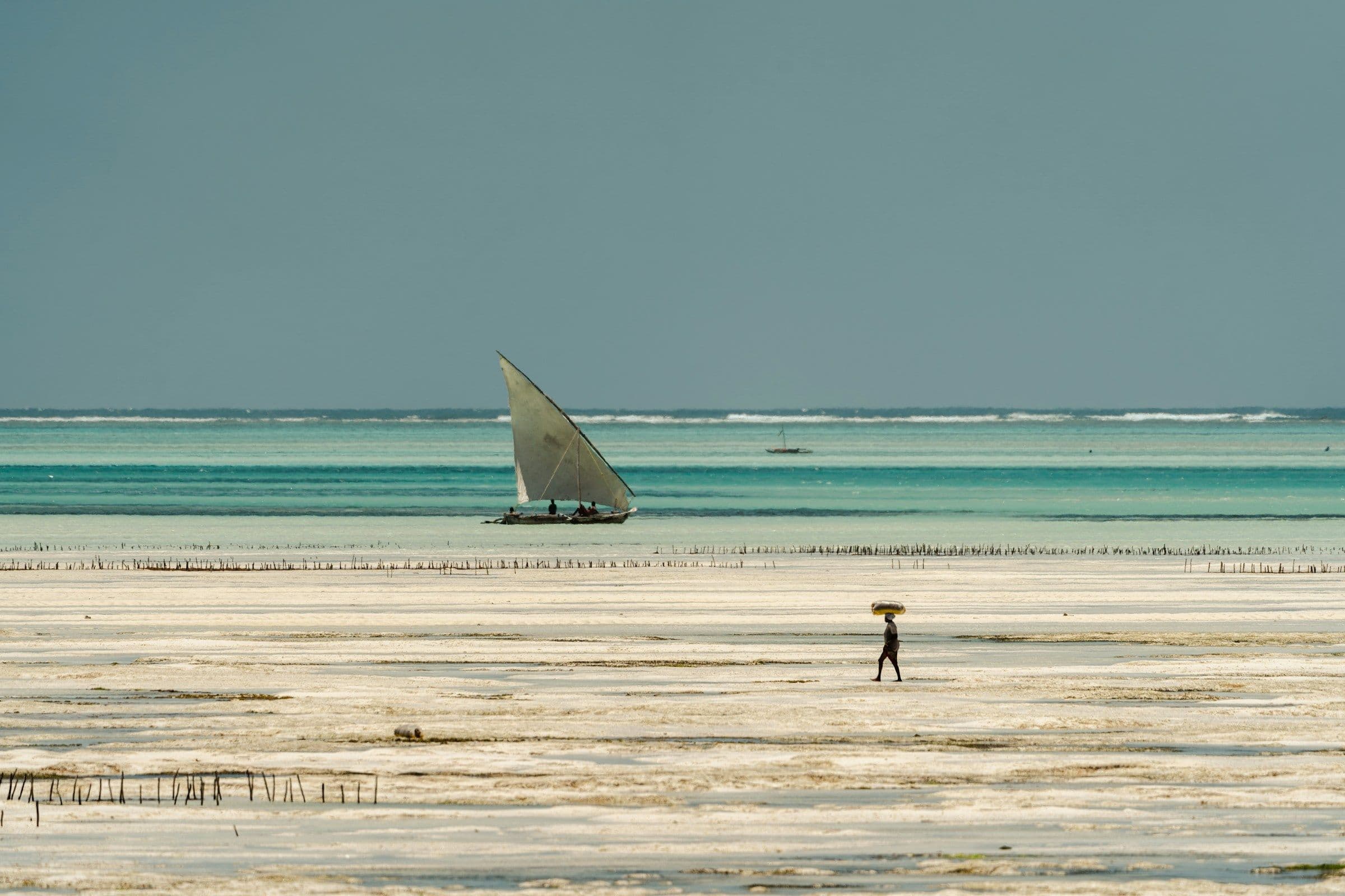 A traditional sailing dhow on turquoise water off a wide white beach at low tide in Zanzibar.