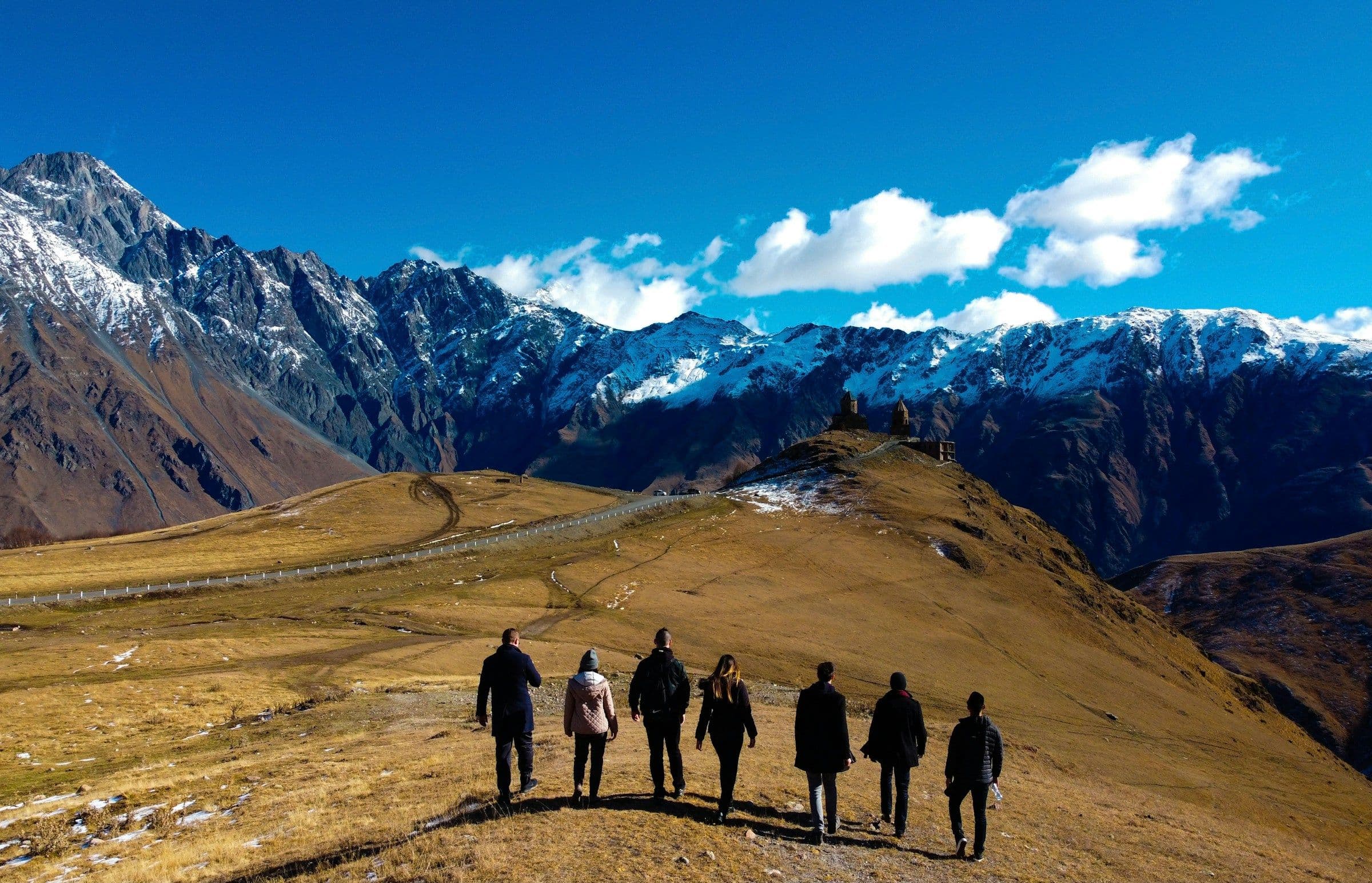 A small group of walkers on a grassy ridge beneath snow-capped mountains in the Caucasus, Georgia.