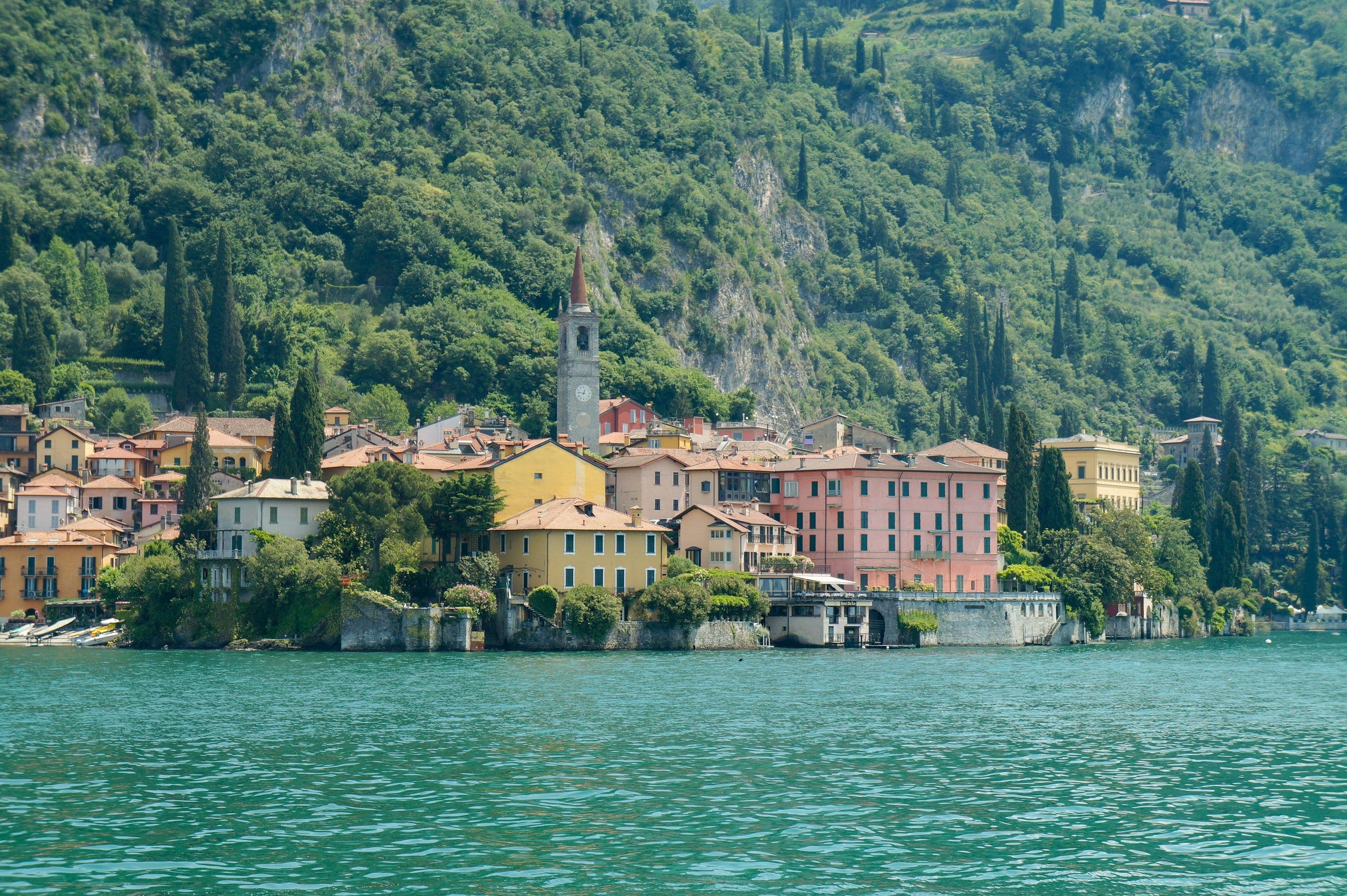 Colourful lakeside houses and a church tower at the foot of a green mountainside on Lake Como, Italy.