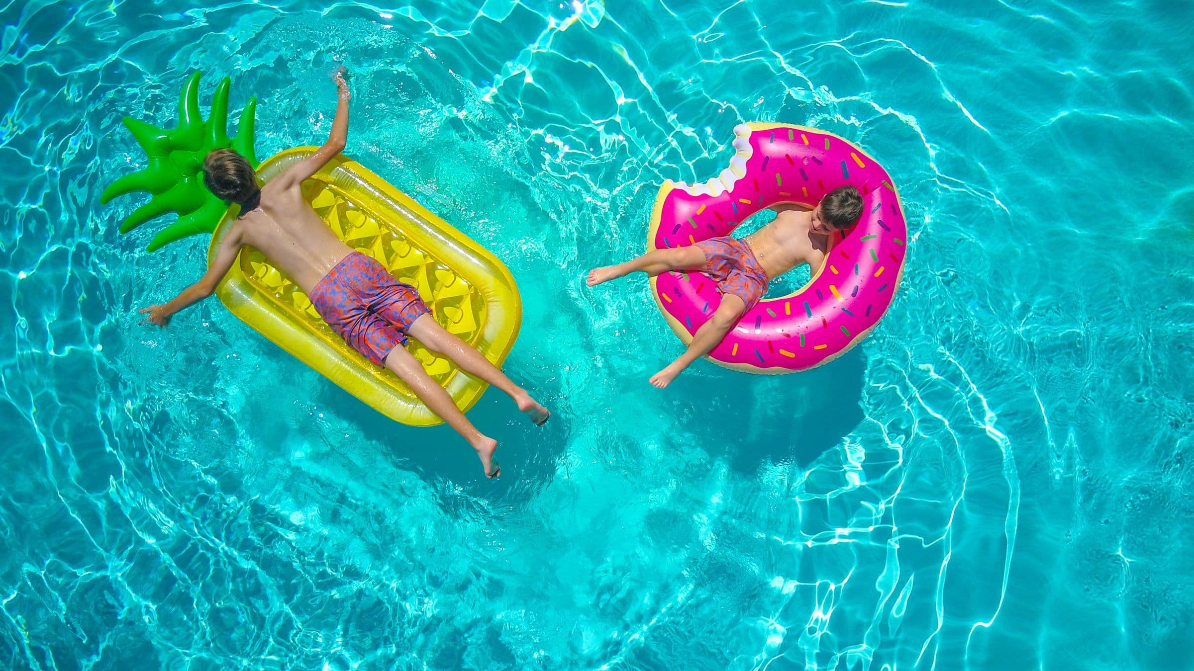 Two children lying on colourful inflatables in a turquoise swimming pool, seen from above.