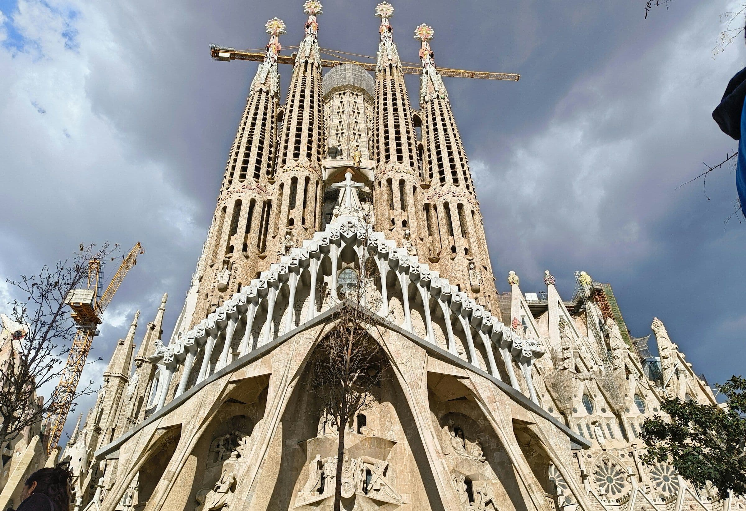 The towers of La Sagrada Família in Barcelona against a cloudy sky, with construction cranes beside them