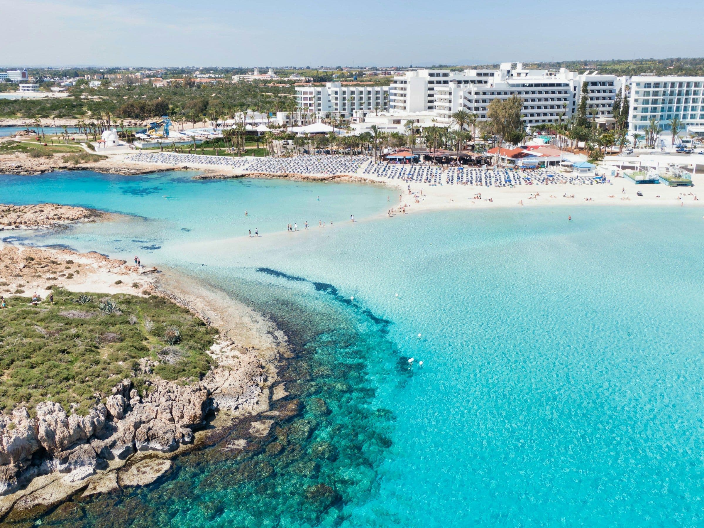 Shallow turquoise water, a sandbar and beachfront hotels at Nissi Beach near Ayia Napa, Cyprus, seen from the air.