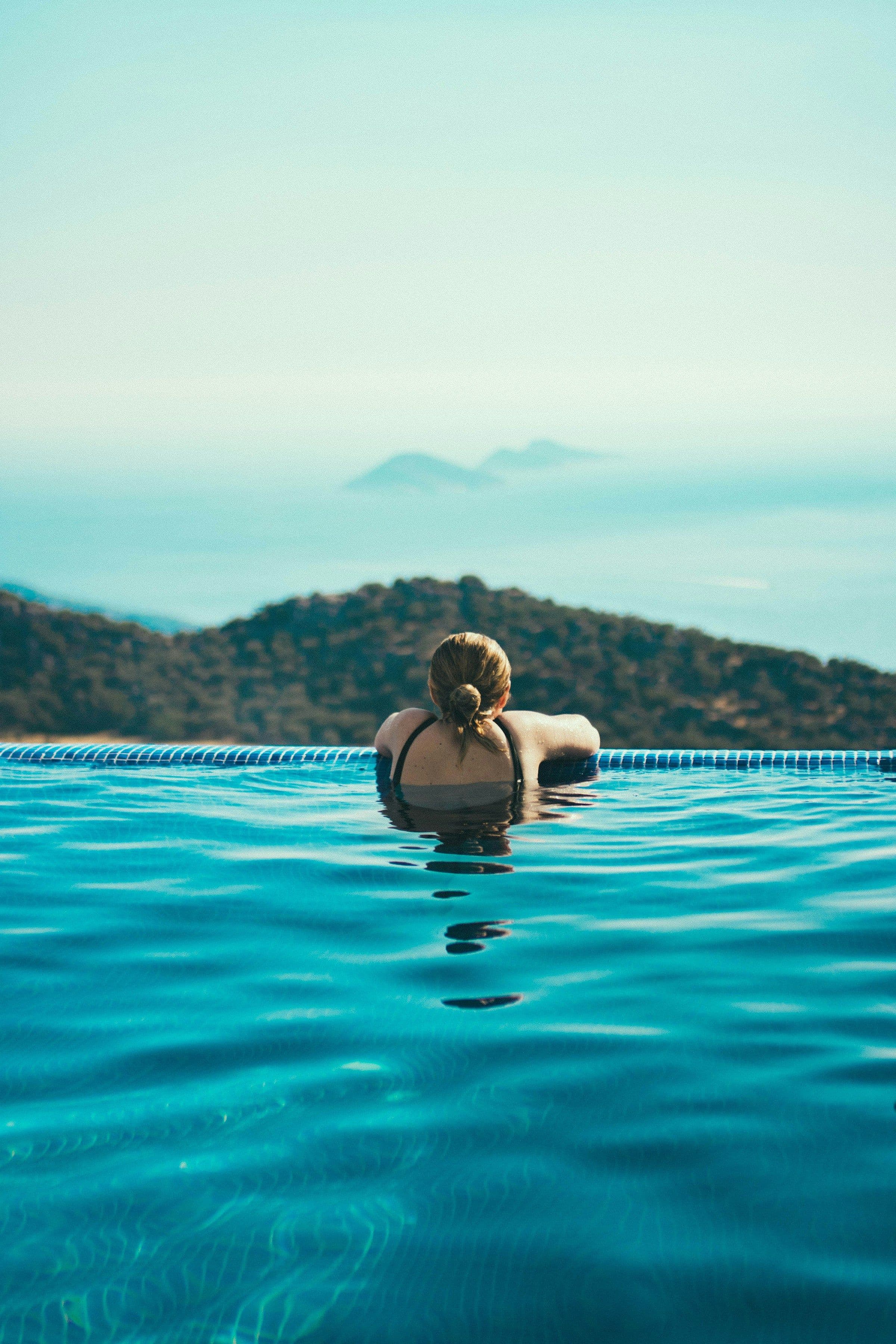 A woman seen from behind, resting at the edge of an infinity pool and looking out to a wooded headland.