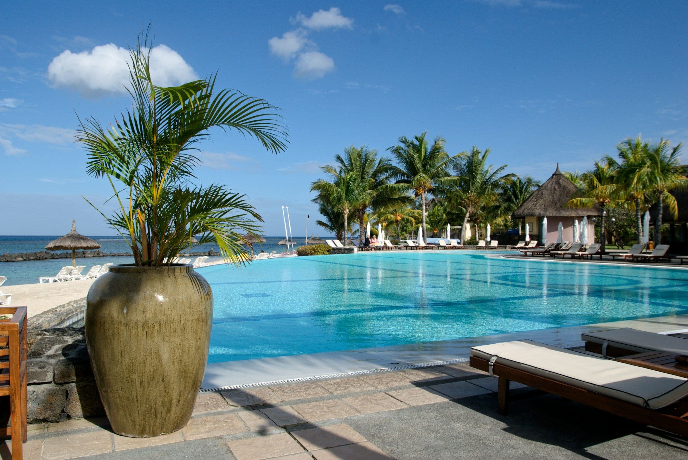 A resort swimming pool edged with palm trees and thatched pavilions beside the sea in Mauritius.