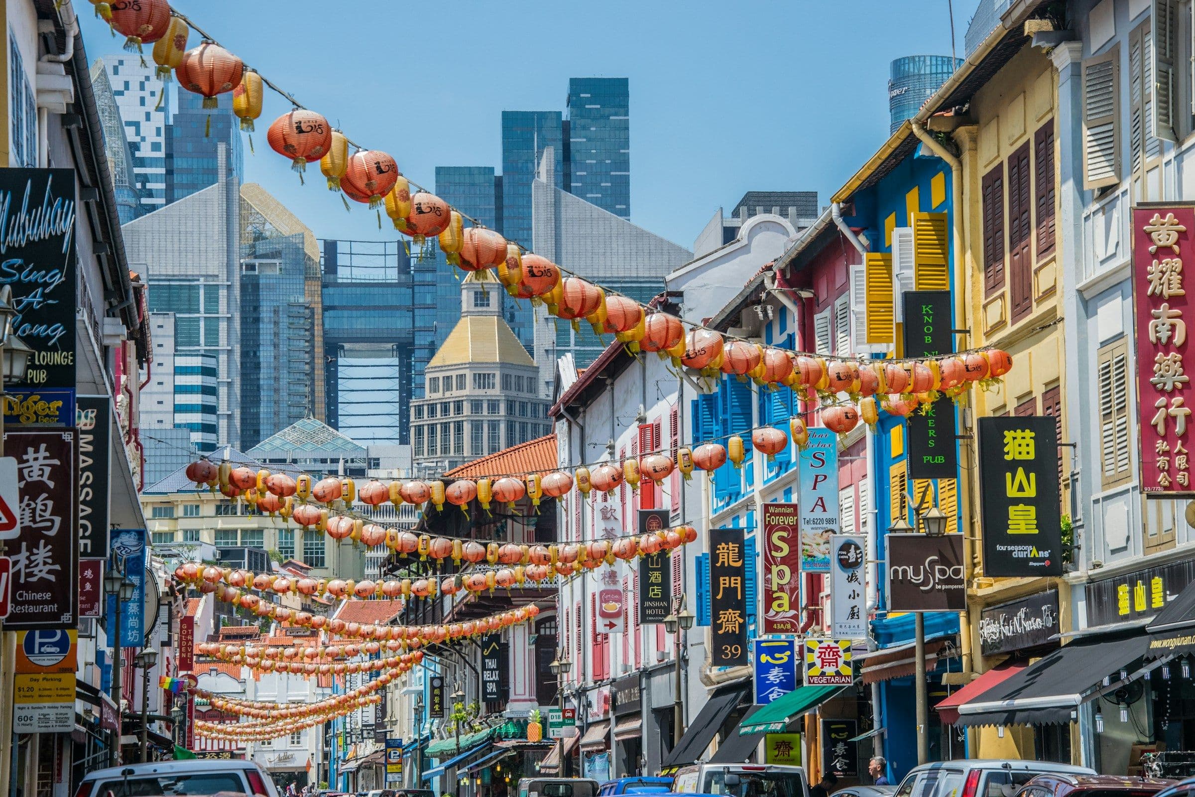 Strings of red lanterns above a street of painted shophouses in Chinatown, Singapore, with modern towers behind.