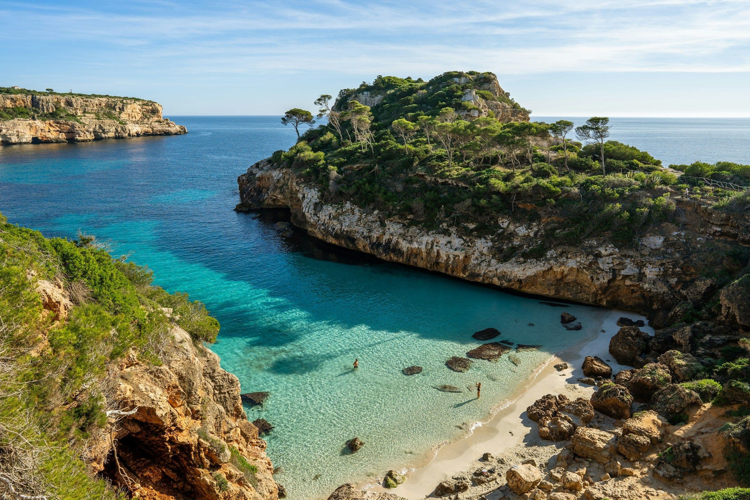 A small sandy cove with clear turquoise water between pine-covered cliffs in Majorca, Spain.