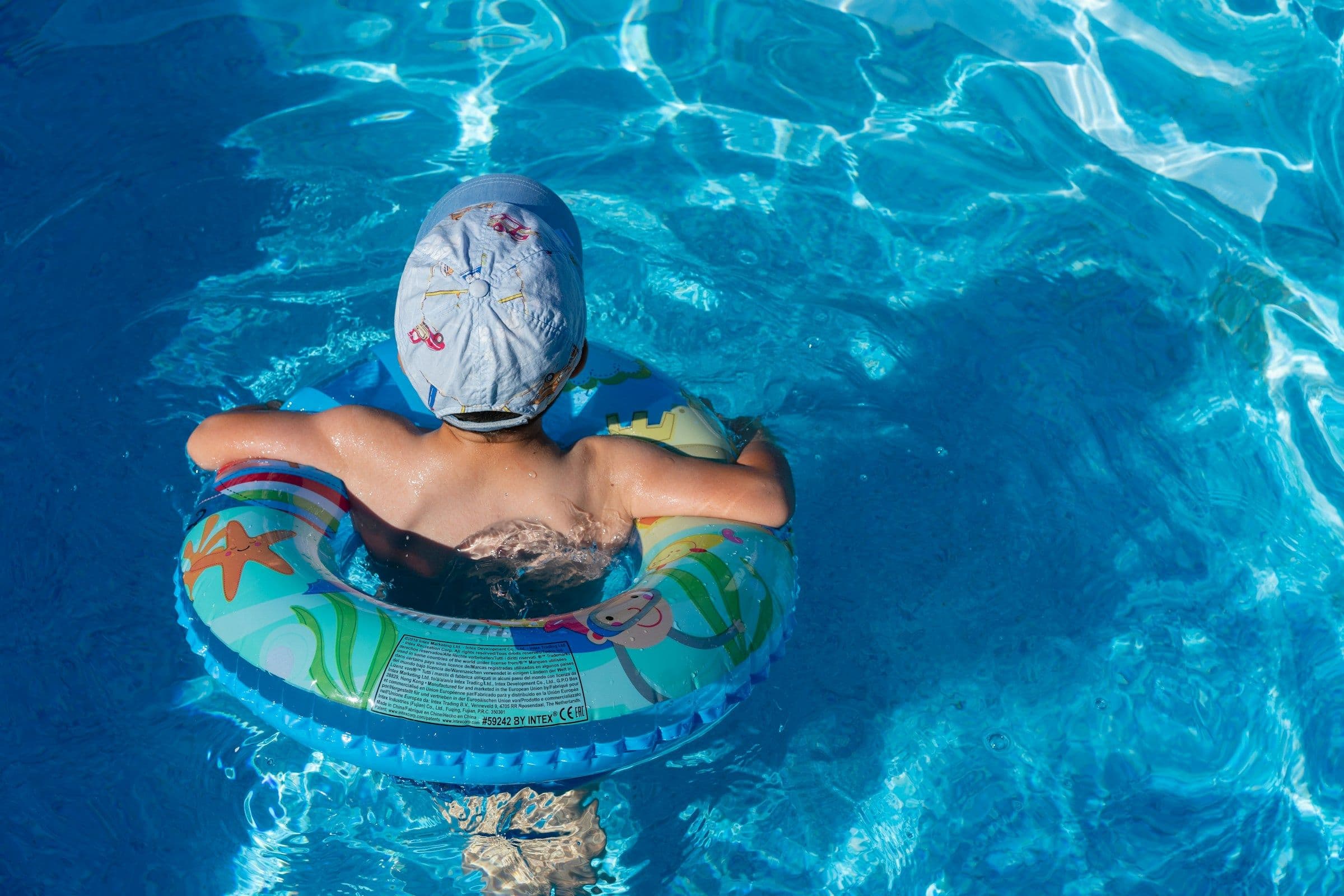 A young child in a sun hat floating in a swim ring in a bright blue pool, seen from above.