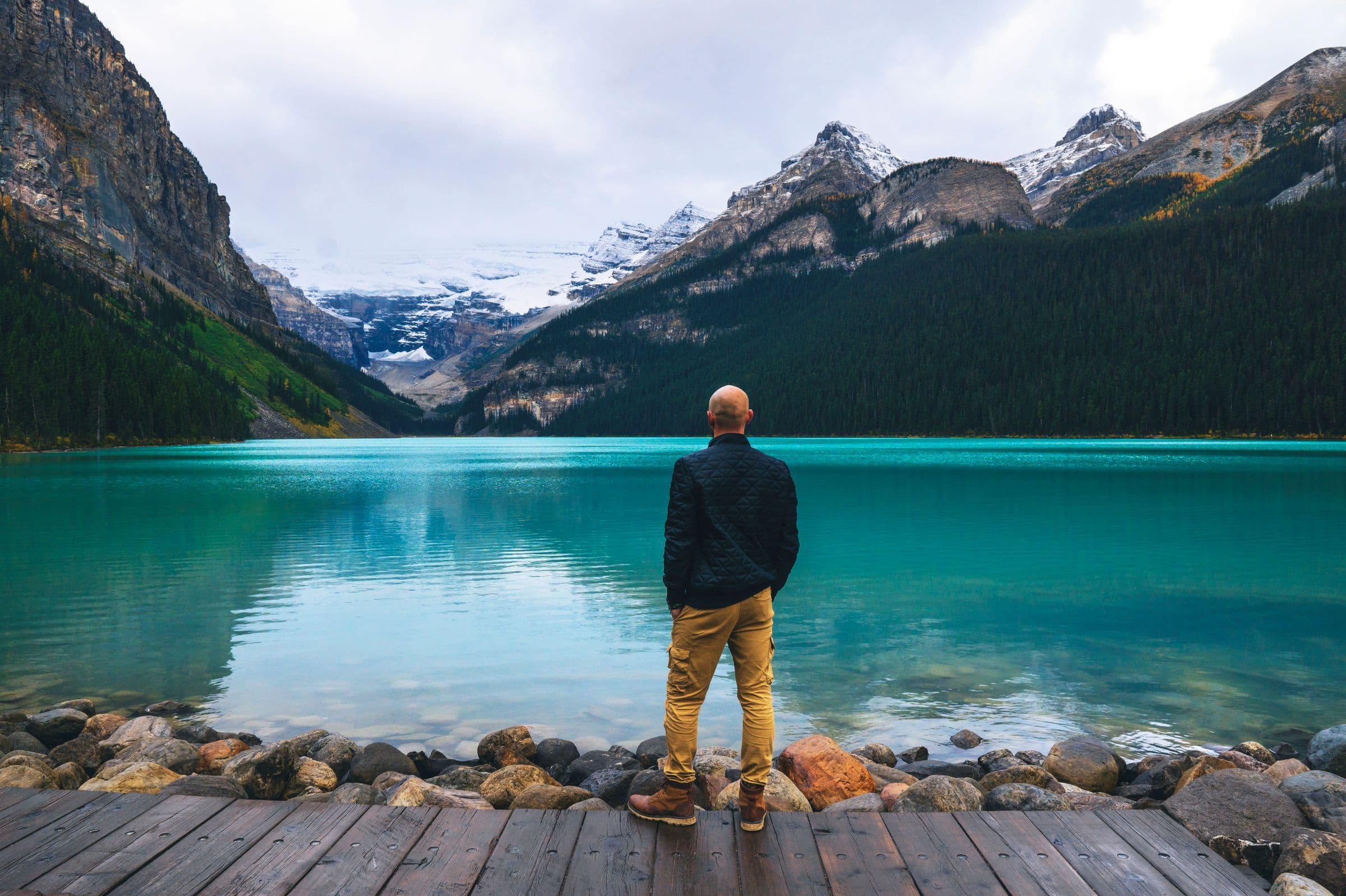 A man seen from behind, standing on a wooden jetty and looking across Lake Louise to the mountains in Canada.