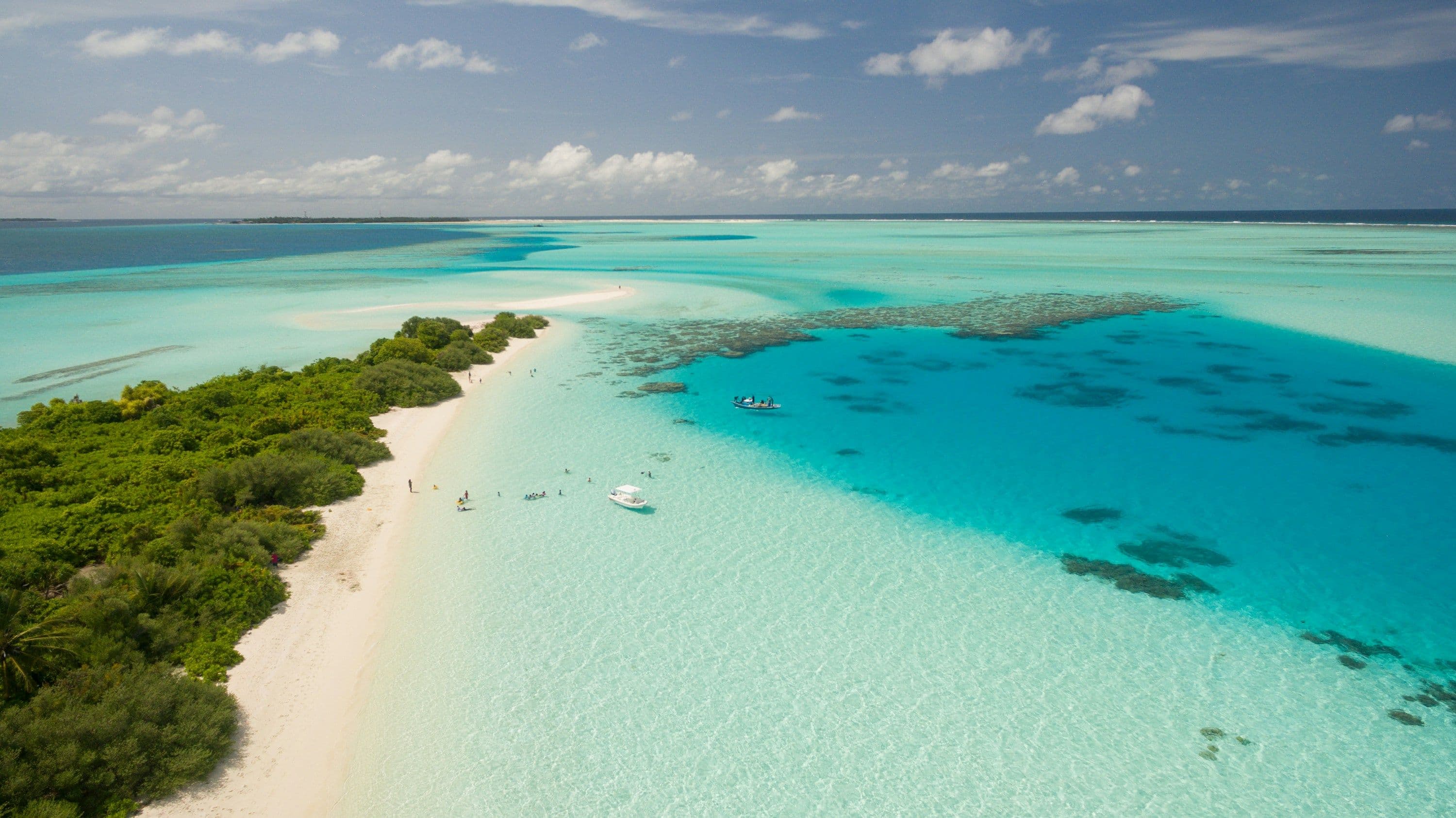 A white-sand beach curving into a turquoise lagoon in the Maldives, seen from the air, with a small boat at anchor.