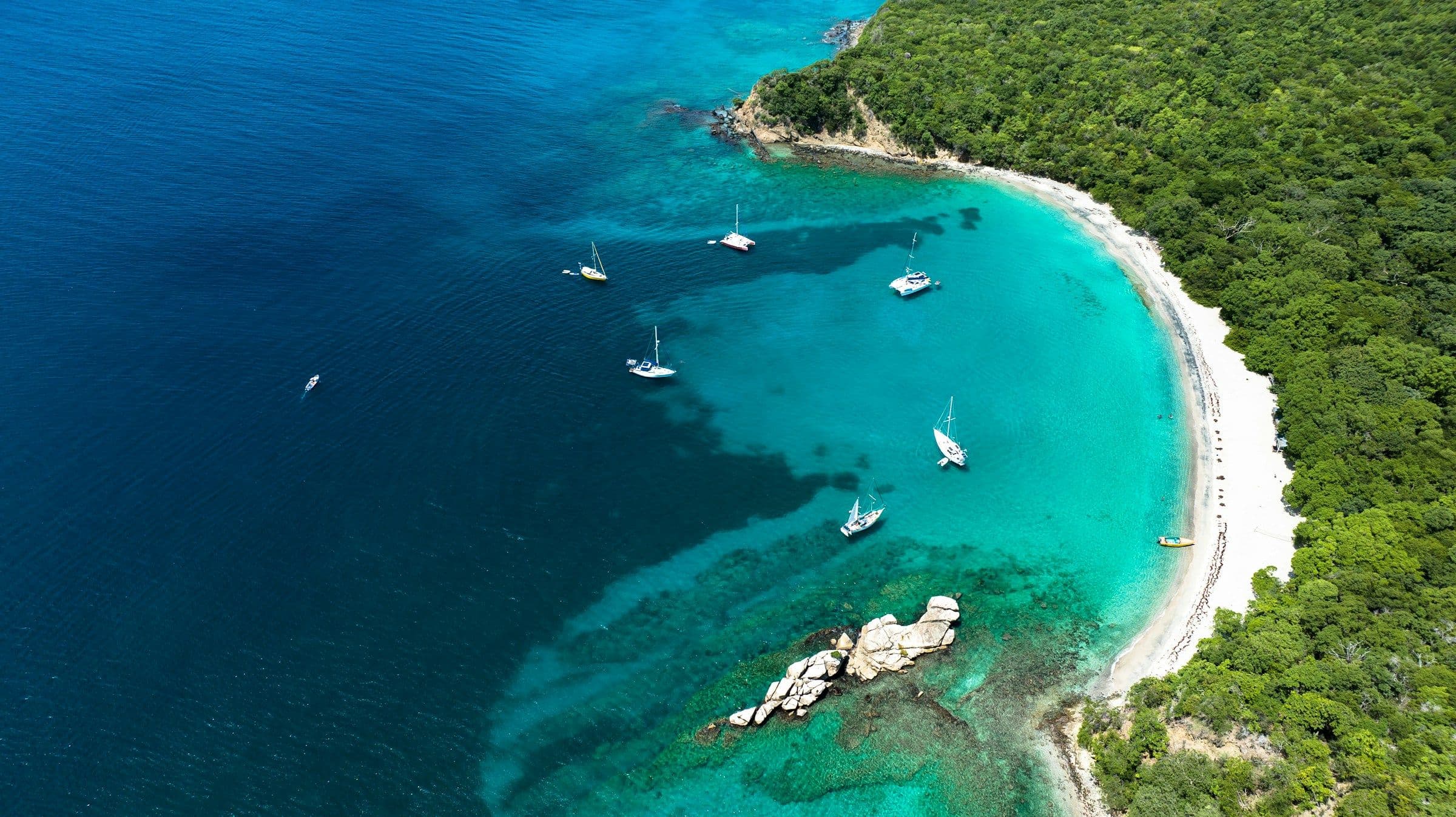 Yachts at anchor in a turquoise bay beside a white-sand beach on the island of Carriacou, Grenada, seen from the air.
