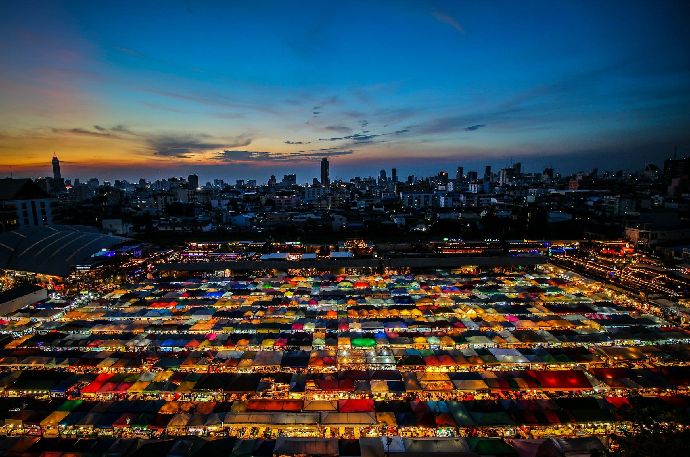 The coloured awnings of a night market in Bangkok, Thailand, seen from above at sunset, with the city skyline behind.