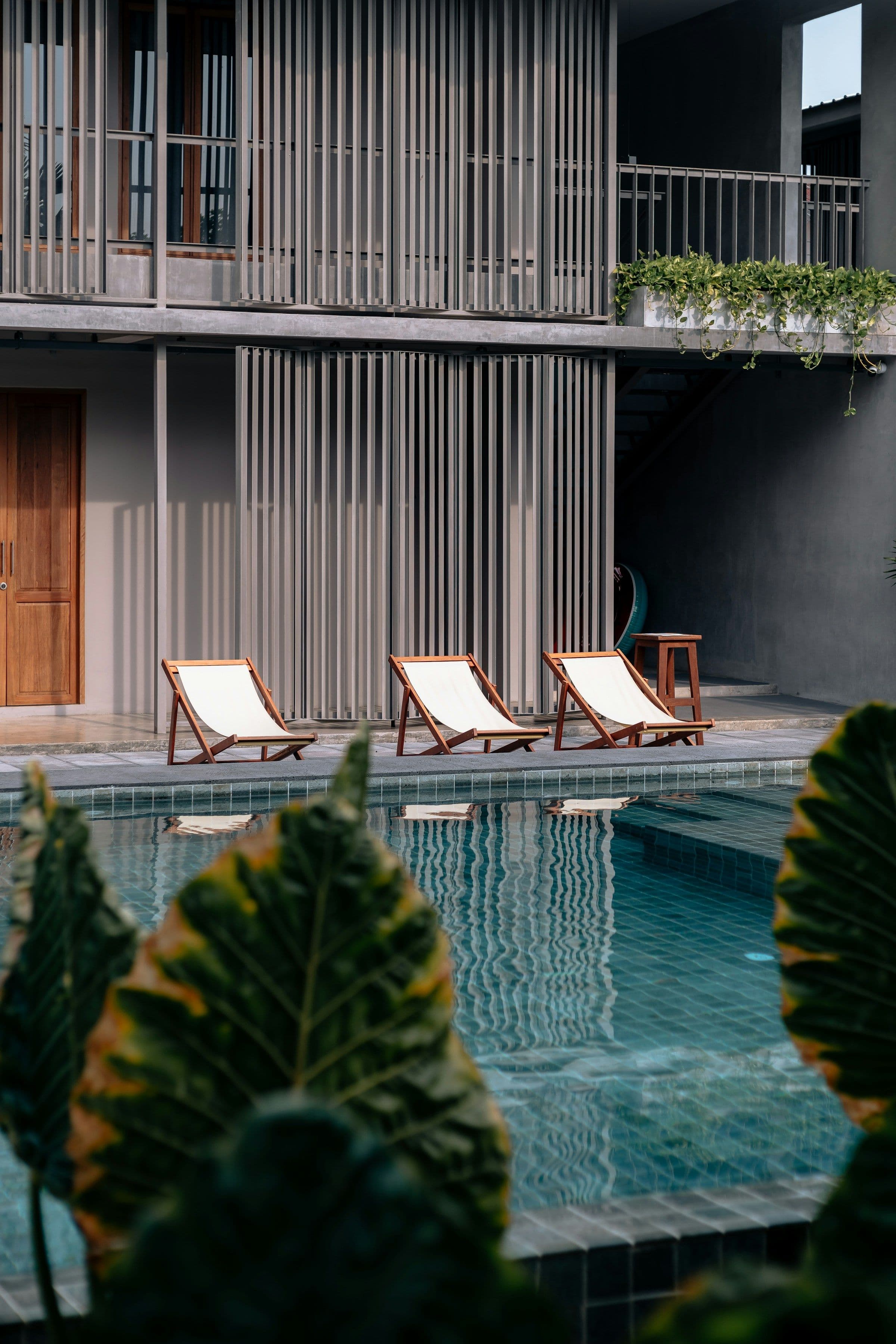 Three wooden sun loungers beside a calm hotel pool surrounded by greenery.