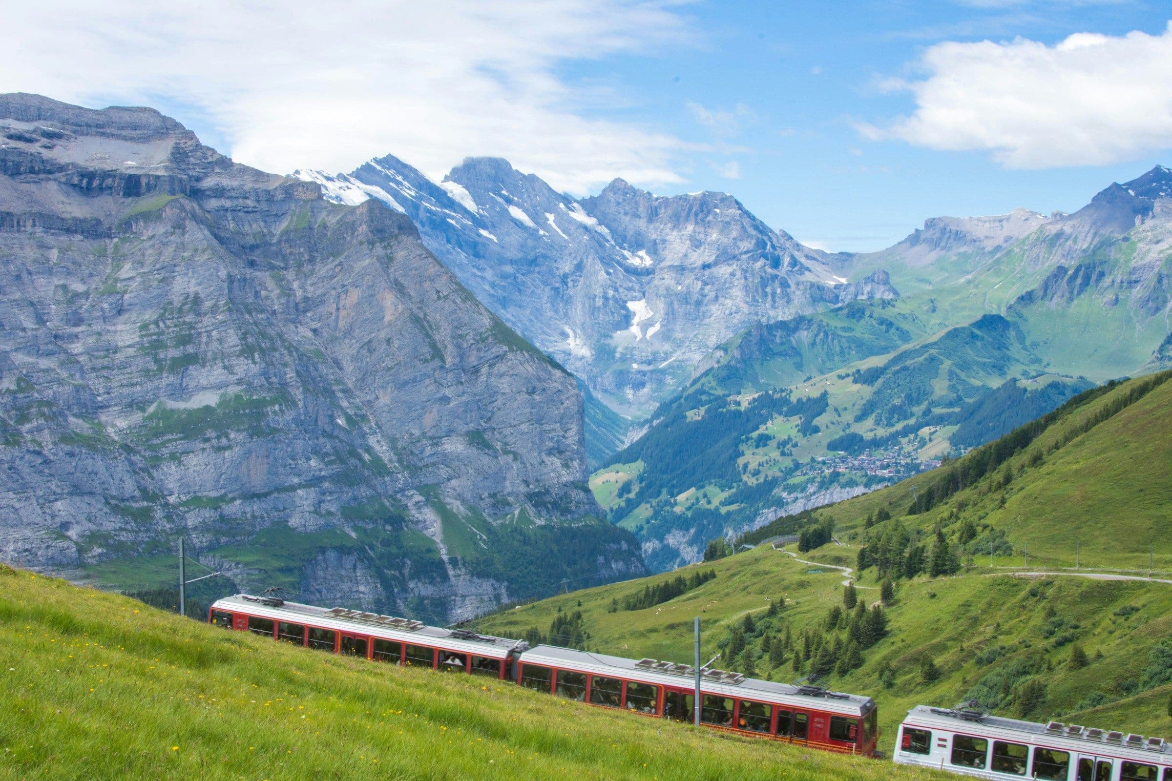 A red and white train crossing green alpine meadows beneath snowy peaks near Lauterbrunnen, Switzerland.