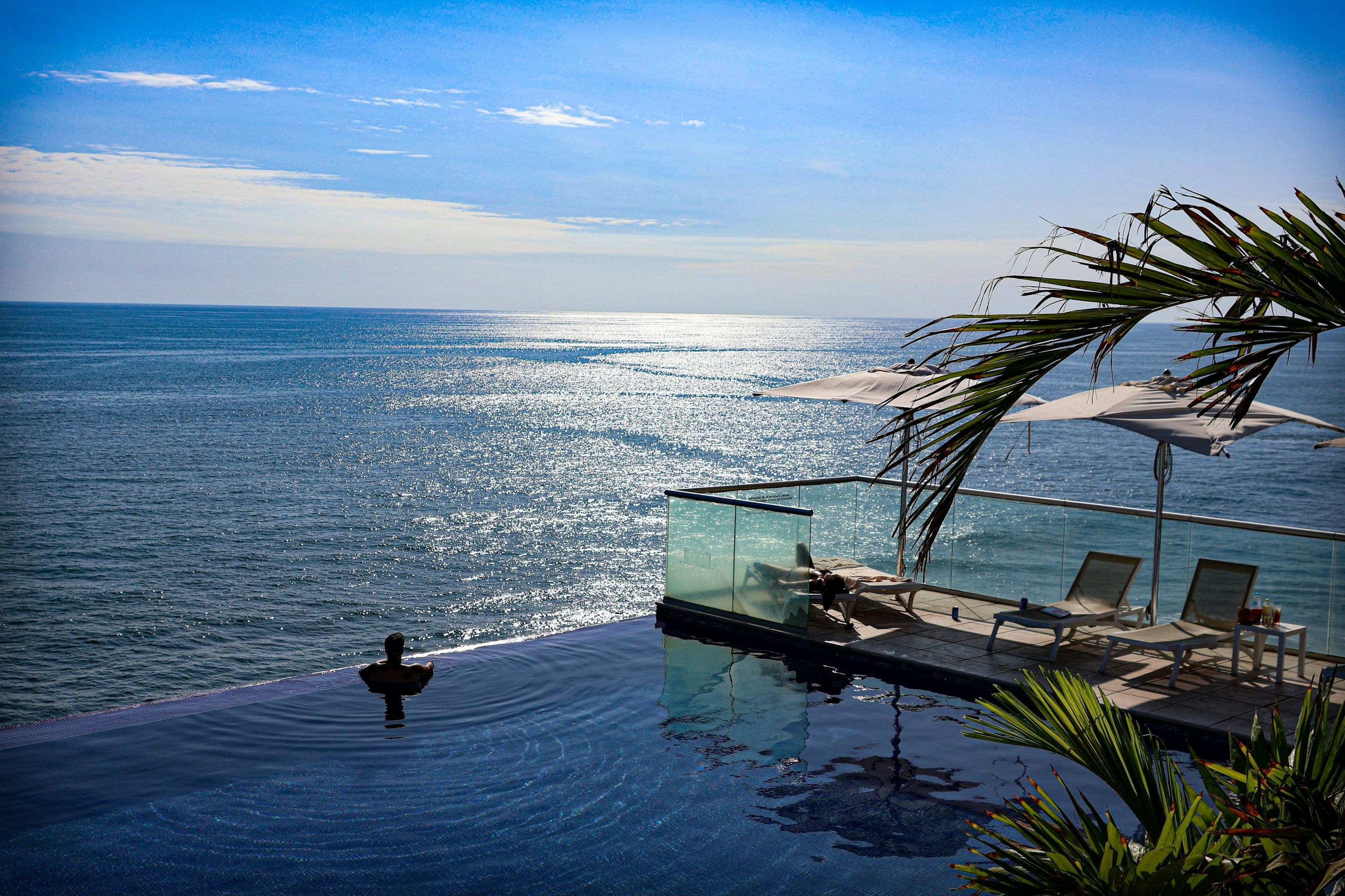 An infinity pool with sun loungers and parasols looking out over the ocean on a clear day.