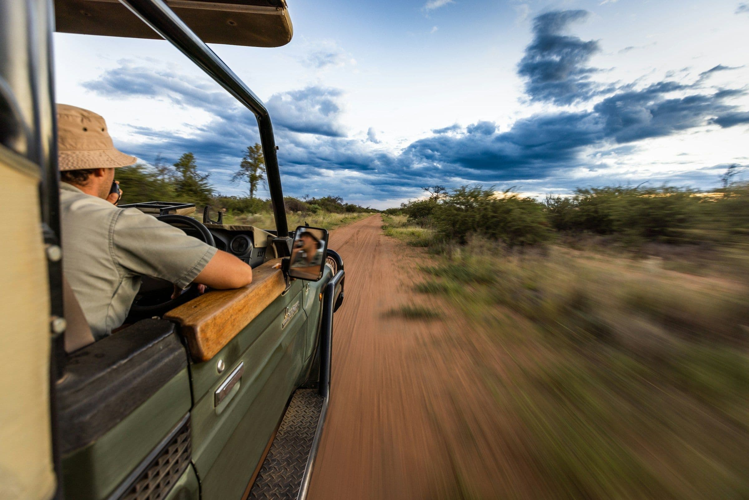 A guide in a hat looking out from an open safari vehicle as it drives along a red dirt track through the bush.