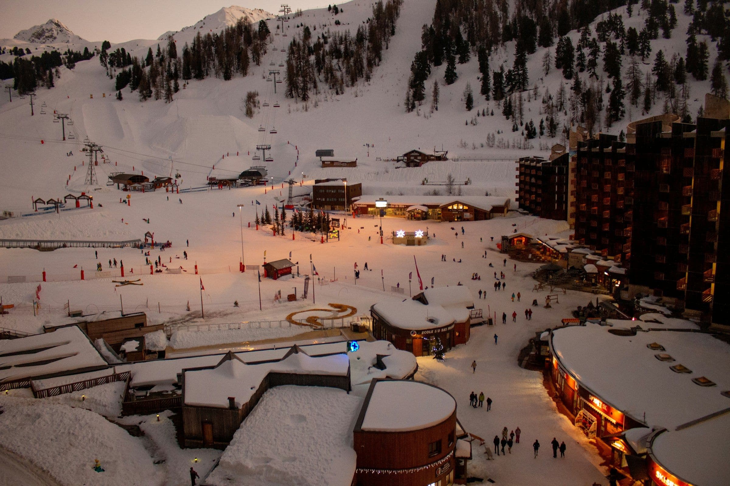 A ski resort village at dusk, its lights coming on among snow-covered buildings and slopes.