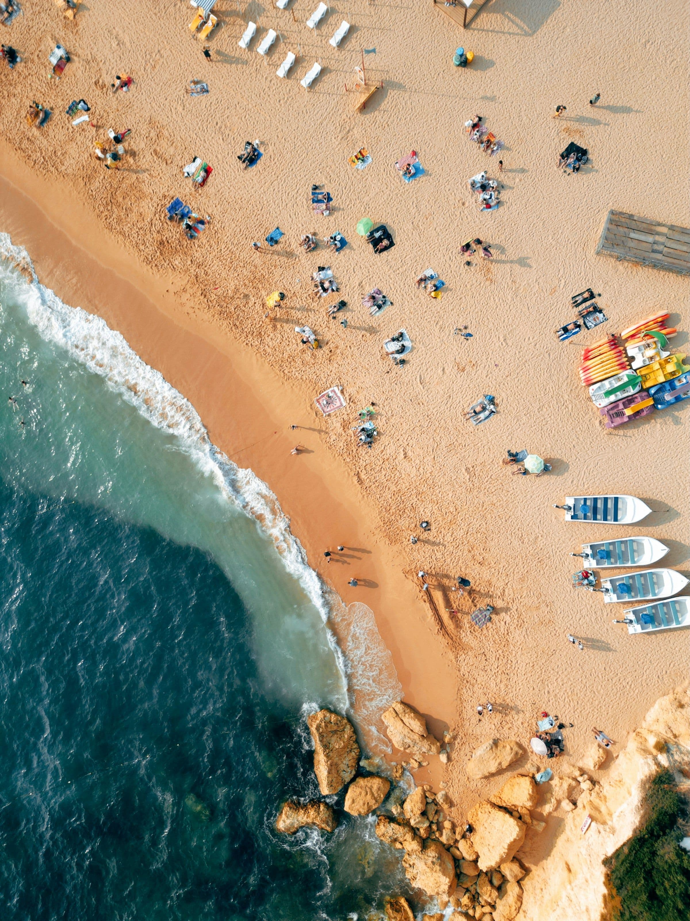 An aerial view of a sandy beach at Carvoeiro, with sunbathers, a row of small boats on the sand and rocks at the water’s edge