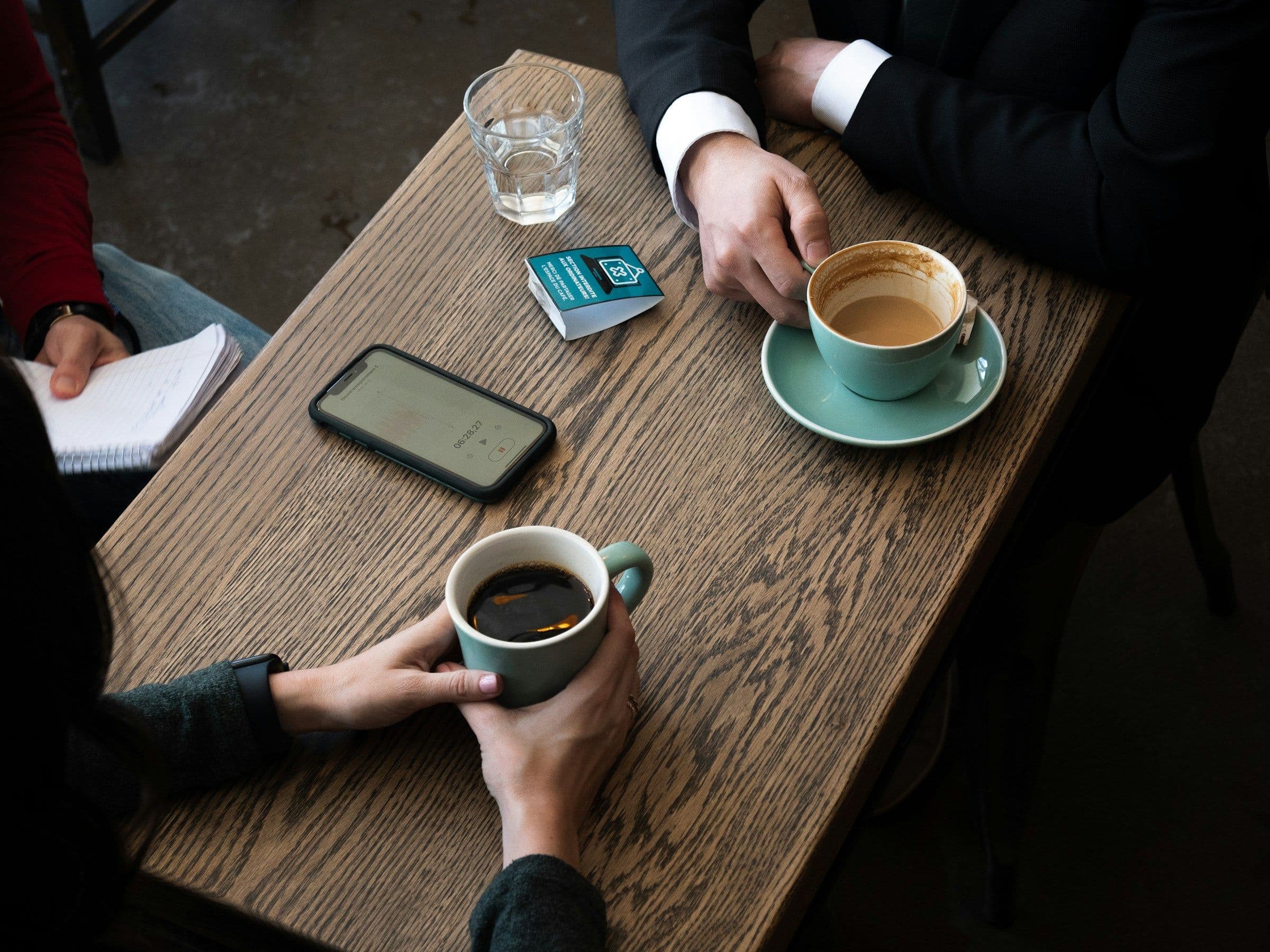 Two people talking over coffee at a wooden café table, one with a notebook open.